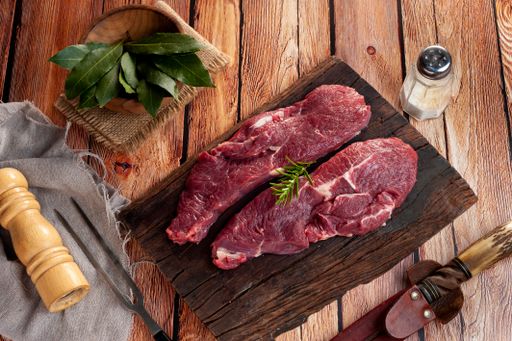A top view of raw striploin beef steaks with rosemary, bay leaves, salt, and  meat fork on the wooden table
