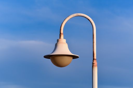 Farola blanca oxidada con bombilla amarilla grande sobre un fondo de cielo azul con unas pocas nubes