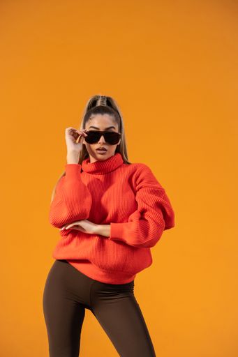 A sexy female in a trendy outfit and black sunglasses is posing in the orange background