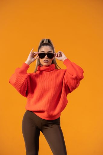 A stylish Spanish model in a warm outfit and sunglasses is standing against the orange background