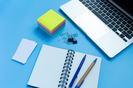 A flat lay workspace with a laptop, a notepad, and two pens, on blue background