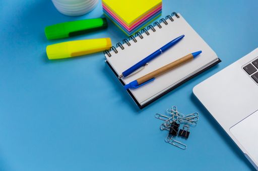 A flat lay workspace with a laptop, a notepad, a and two pens, on blue background
