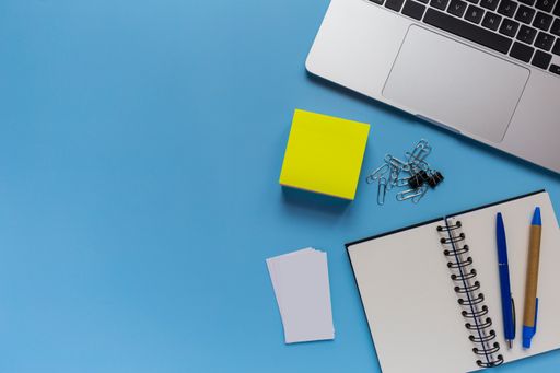 A flat lay workspace with a laptop, a notepad, and two pens, on blue background