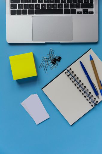 A flat lay workspace with a laptop, a notepad, and two pens, on blue background