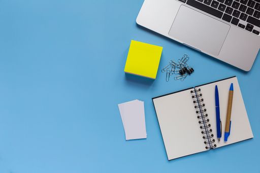 A flat lay workspace with a laptop, a notepad, and two pens, on blue background