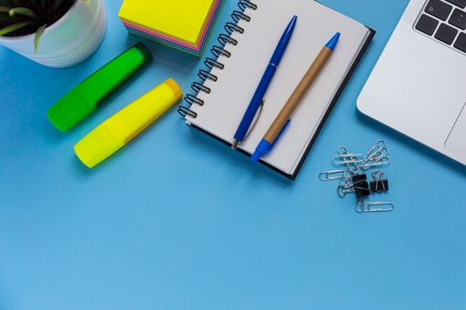 A flat lay workspace with a laptop, a notepad, and two pens, on blue background