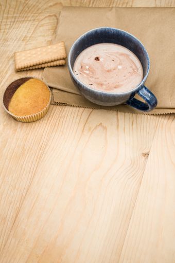 hot milk with cocoa in a blue cup atasty cupcake and biscuits on a rustic wooden and brown paper background