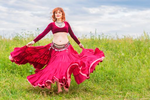 Young beautiful red-haired girl in a red dress enthusiastically dances a fascinating dance on a green lawn under the open blue sky.