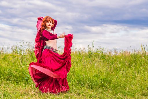 Young beautiful red-haired girl in a red dress enthusiastically dances a fascinating dance on a green lawn under the open blue sky.