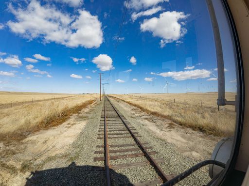 View from a train window showing a railway track stretching through a dry landscape with wind turbines and a bright blue sky.