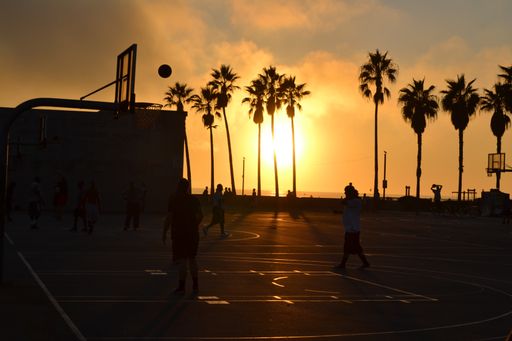 A silhouette shot of people playing basketball at a basketball court near coconut trees during sunset