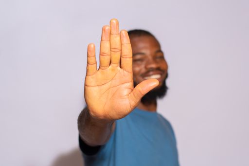 A black man with a blue t-shirt smiling and holding his palm out with a white background