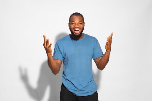 A black man with a blue t-shirt cheerfully celebrating a win with hands up on a white background.
