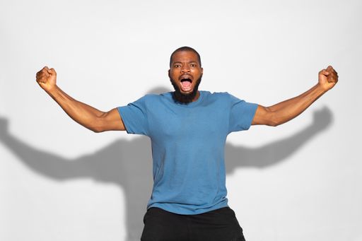 A black man with a blue t-shirt cheerfully celebrating a win with fists up on a white background.