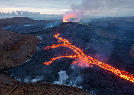 A long lava river going over hills and fields with a background of an erupting volcano