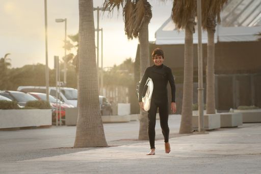 A smiling and happy teenage surfer holding his surfboard standing next to palm trees.