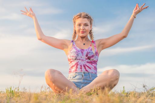 Young beautiful and tanned girl sits in lotus position on grass with her hands raised up against background of blue summer sky. Looks into camera