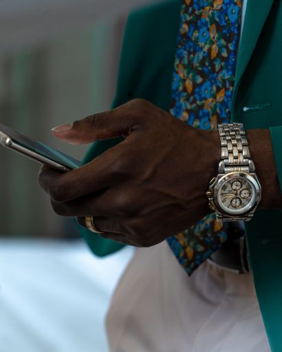 A vertical closeup shot of an African male wearing green suit and a gray wristwatch holding a smartphone