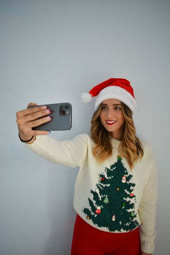 A Caucasian female taking a selfie in a Santa Claus hat and a sweater with a Christmas tree