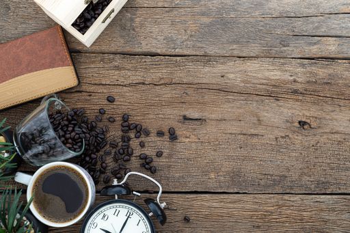 A top view of a wooden desk with coffee mug, beans, alarm clock and notebook