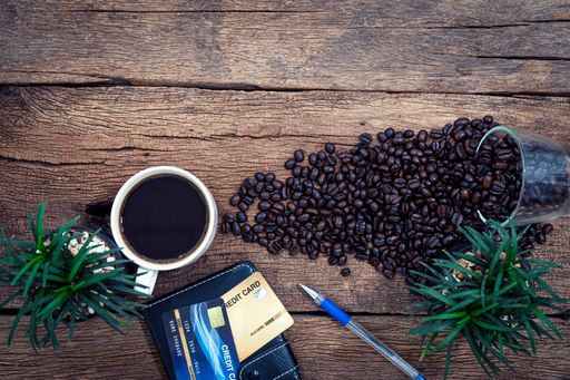 A top view of a work desk with a cup of coffee, roasted coffee beans, a wallet and plants