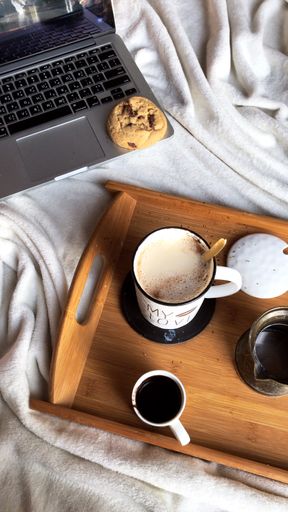 A vertical shot of a cup of homemade coffee on a wooden tray on a cozy blanket next to a laptop