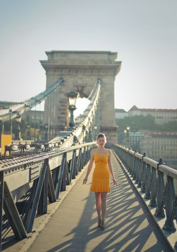 A vertical shot of a female in a yellow dress walking through the Szechenyi Chain Bridge in Budapest