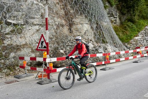 An elderly woman cycling on a road in the Switzerland countryside with rockfall netting on a cliff beside it