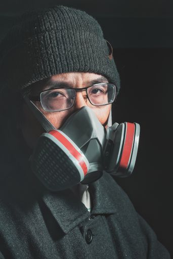 A portrait of an east asian male with glasses wearing a respirator on a dark background