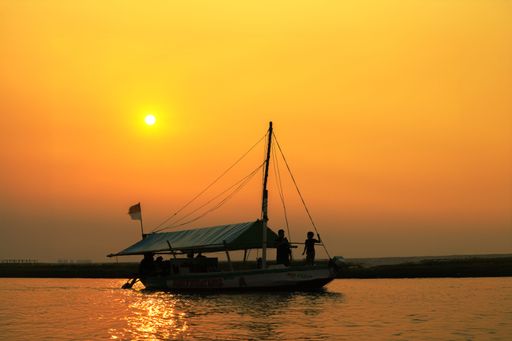 A silhouette of a small fishing boat sailing under golden sky at sunset, creating warm reflections on the water.