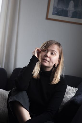 A young blonde woman wearing a black turtleneck sweater sitting in a sofa looking into the camera.