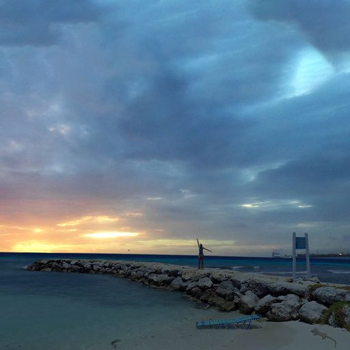 A person stands on a rocky pier at sunset, with a dramatic sky and calm sea in the background.