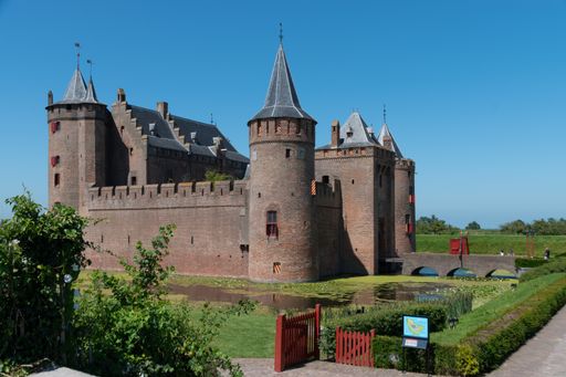 Side view of the Muiderslot. The Muiden castle is surrounded by water, the bridge acts as a passageway and many green plants surround the area.