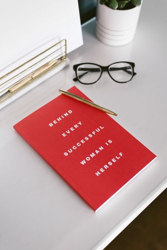 A closeup view of a white desk with white papers, red notebook, plant in a white bowl, pen and glasses in it