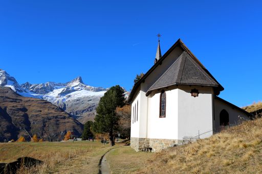 Charming alpine chapel near Zermatt, Switzerland, with snowy mountain peaks and blue sky in the background.