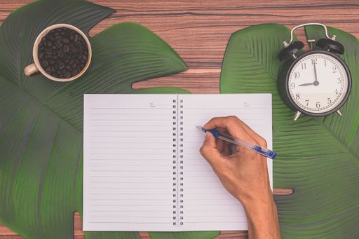A top view of hand holding pen on a notebook with alarm clock, and a mug in a desk