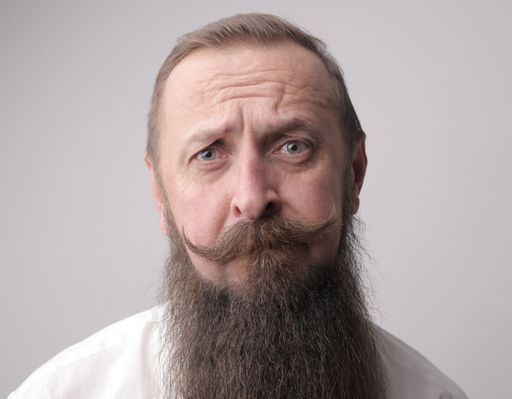 A man with a long beard and a mustache frowning while standing in front of a grey wall