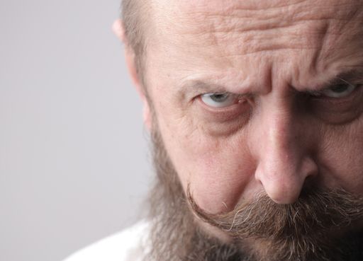 A man with a beard and a mustache frowning while standing in front of a grey wall