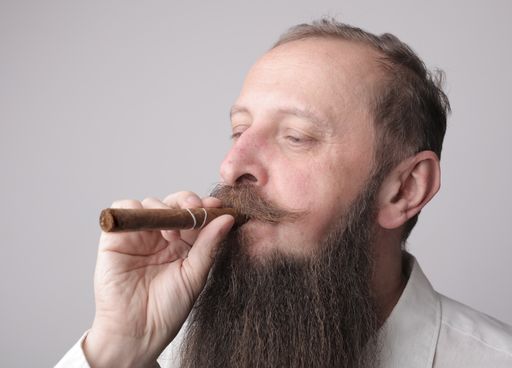 A man with a long beard and a mustache smoking a cigar with a grey wall on the background