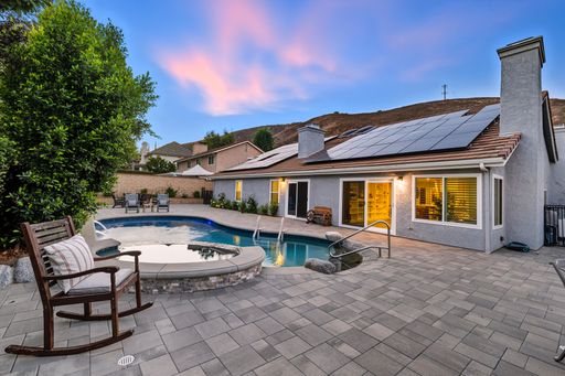 Outdoor living space with pavers, built-in fireplace, dining area, and spa surrounded by hedges at sunset