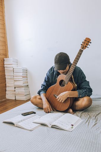 Young man practicing guitar with sheet music in cozy room, surrounded by books, wearing casual clothing, sitting on a bed.