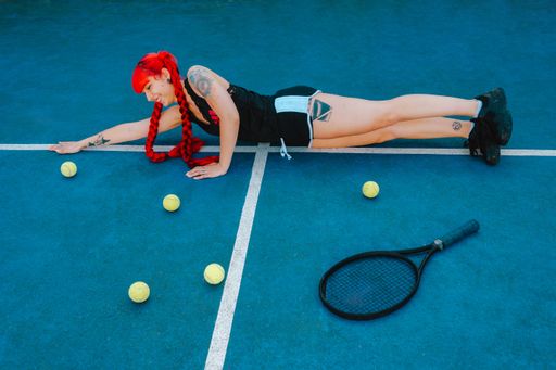 A young woman with red hair laying on the tennis court with a racket near her