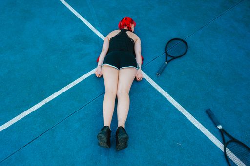 A young woman with red hair laying on the tennis court with a racket near her