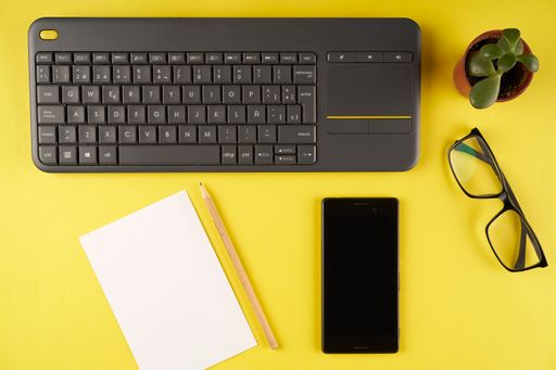 A flat lay shot of keyboard, smartphone, glasses, notepad and pencil on a yellow background