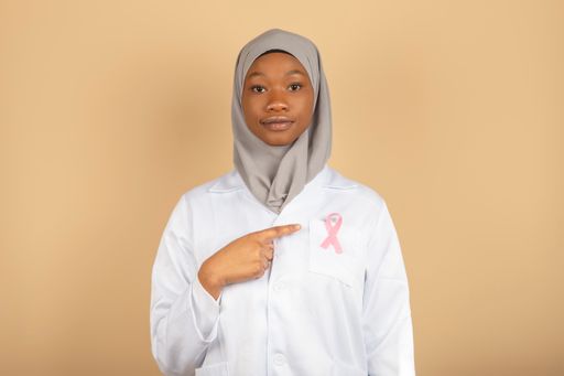 An African female model in a doctor uniform pointing to a pink breast cancer awareness ribbon.