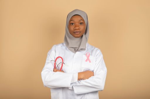 An African female model in a doctor uniform with a pink breast cancer awareness ribbon and arms crossed.