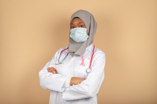 An African female model in a doctor uniform with a pink breast cancer awareness ribbon and arms crossed.