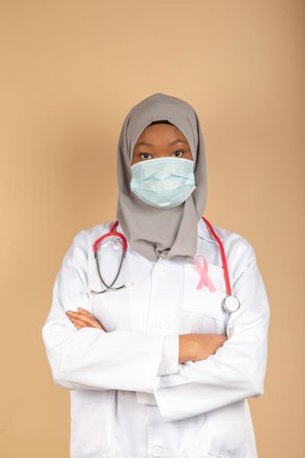 A vertical shot of an African female model in a doctor uniform with a pink breast cancer awareness ribbon and arms crossed.