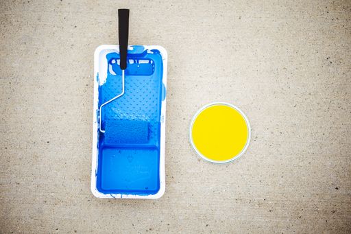 A top view of a paint roller in tray with blue paint and a can of yellow paint on a concrete ground