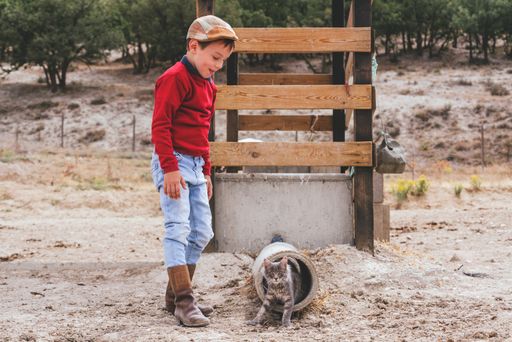 A Caucasian village boy wearing boots and a hat with a kitten on a farm